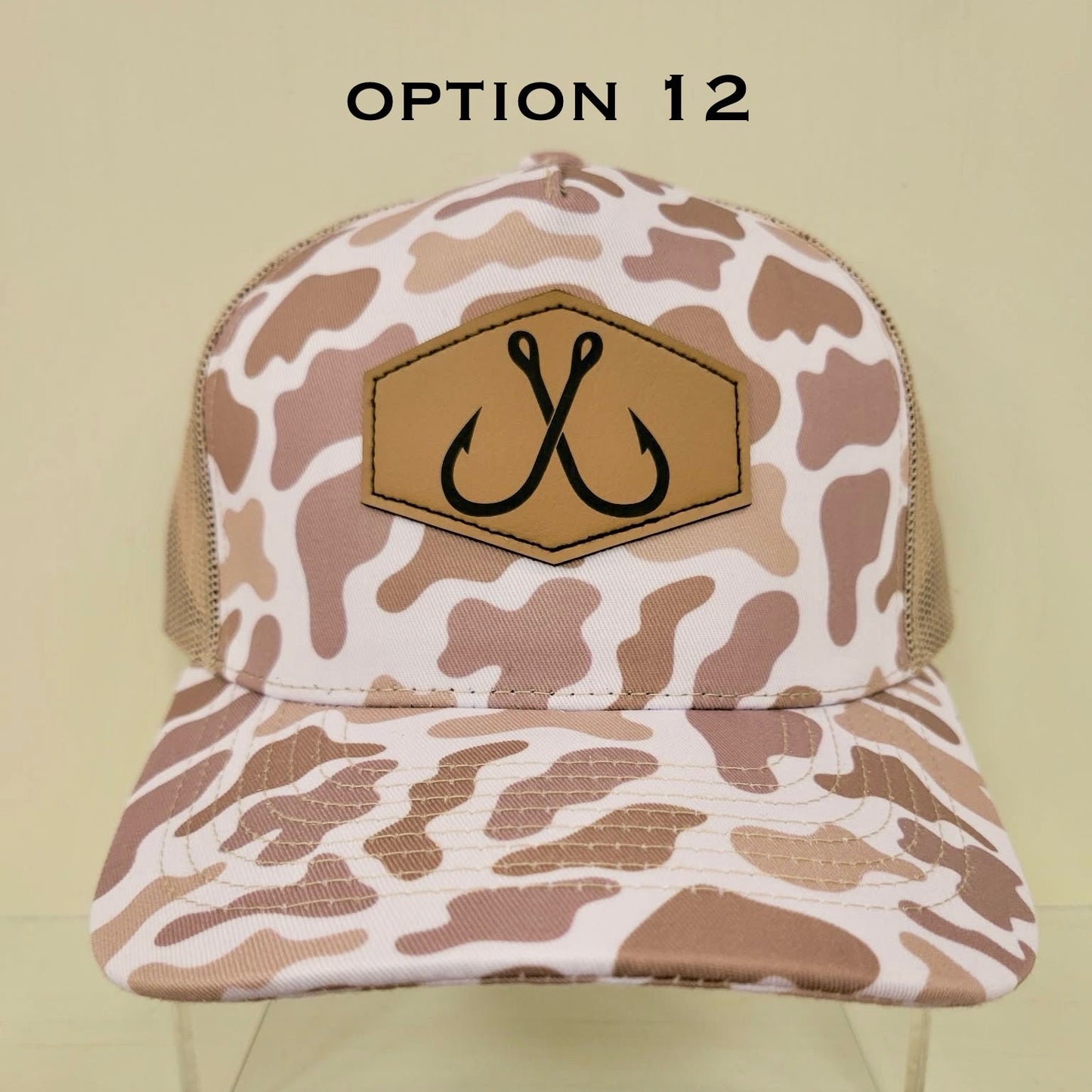 Camouflage-patterned cap with a patch featuring two fishhooks on a beige background.