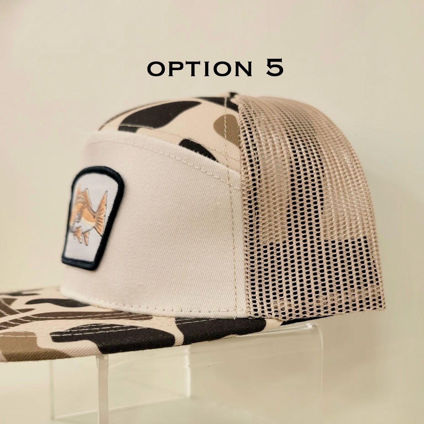 Beige trucker cap with camouflage pattern and patch on a clear stand against a light background